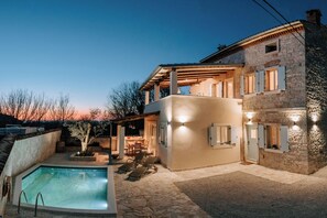 Una casa de piedra de dos plantas con piscina, mobiliario de exterior y un porche cubierto.