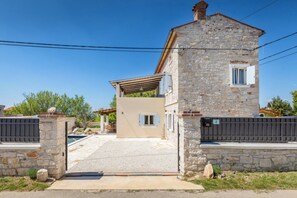 Una casa de piedra con un porche cubierto, un balcón pequeño y un camino de entrada cerrado por una reja.