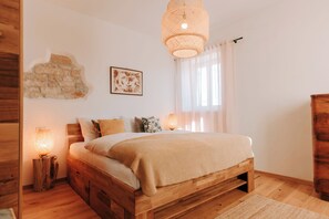 Uma cama de madeira com cabeceira, uma luminária na parede, um quadro emoldurado e uma janela com cortinas.