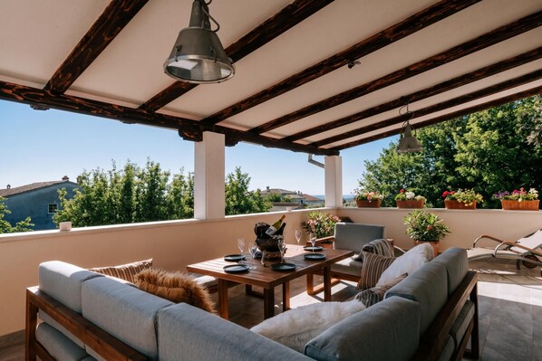 Un área de estar al aire libre cubierta, con mesa lista para comer, asientos acolchados y plantas en macetas.