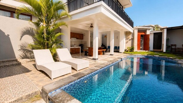 Un área de piscina moderna con sillones de descanso, un porche cubierto y una piscina azul cristalina.