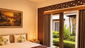 Un dormitorio con una cama grande, un cuadro de paisaje tropical, una mesita de noche y vistas a un jardín a través de una puerta corrediza.