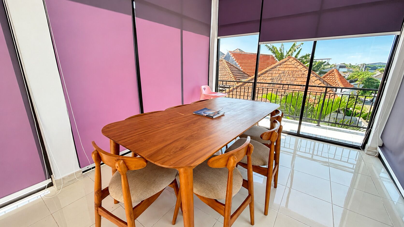 A room with a wooden dining table, four chairs, a window with a view of houses and greenery, and purple walls.