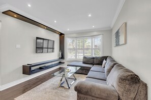 A modern living room with a sectional sofa, a glass coffee table, a mounted TV, and a built-in bench with storage. - Modern 4BR Family Home w/ Pool · Whitby (Whitby)