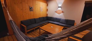 A room with a wooden floor, a dark-colored sofa, a wooden table, and a hammock. - New villa for 8 people • Large covered terrace • 10 minutes beaches of Soulac (Talais)