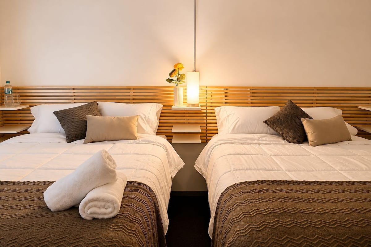 Hotellihuone, jossa on kaksi sänkyä, yöpöytä lampun kanssa ja seinälle kiinnitetty hylly.