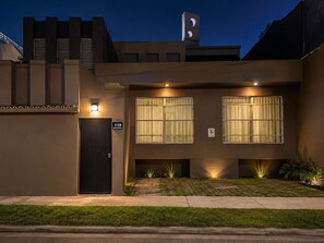 A modern building with a black door, two windows with barred glass, and a small garden area with plants.