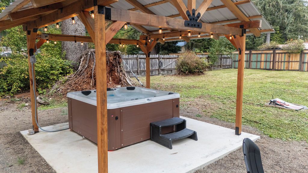 A hot tub under a wooden pergola with string lights, surrounded by a backyard with a wooden fence and greenery.