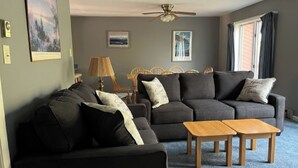 A living room with a sofa, two wooden side tables, a ceiling fan, and a window with curtains. - Open Pit by Avantstay Indoor Heated Pool + Rec Center Near Gore + Hudson (North Creek)