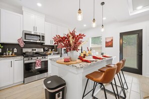 A modern kitchen with white cabinets, stainless steel appliances, a central island with bar stools, pendant lighting, and a decorative plant. - Near Med Center and NRG Stadium, Game Room, Fireplace (Houston)