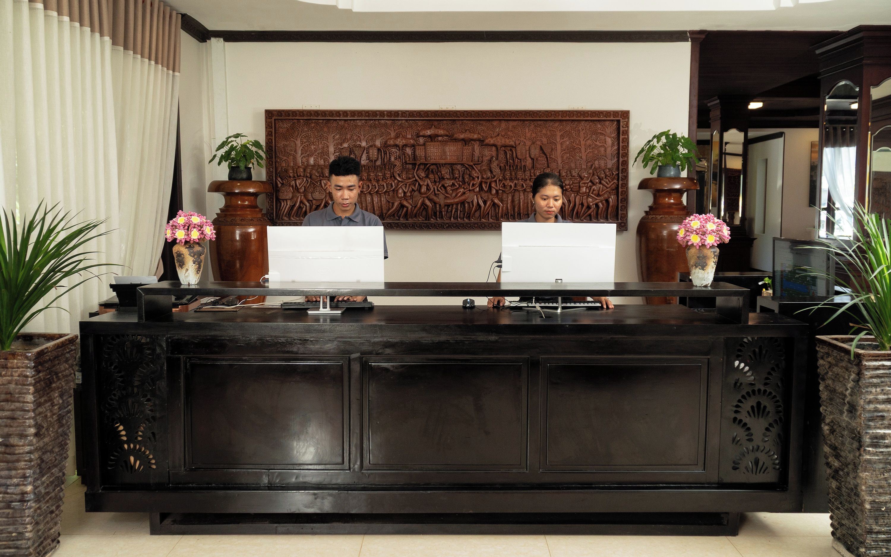 A reception desk with two individuals seated behind it, a decorative wall piece above, and potted plants on either side.