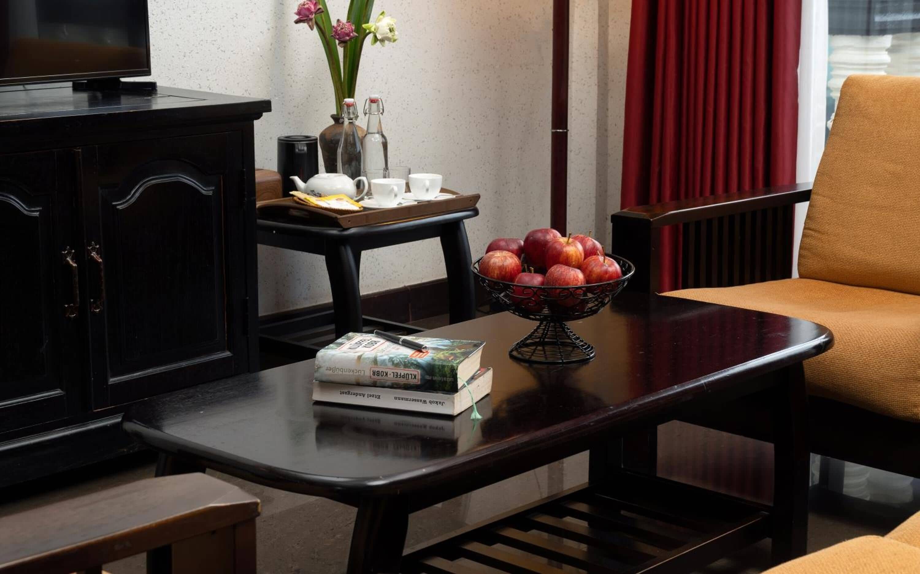 A room with a dark wooden cabinet, a table with books, a bowl of apples, and a chair with a cushion.
