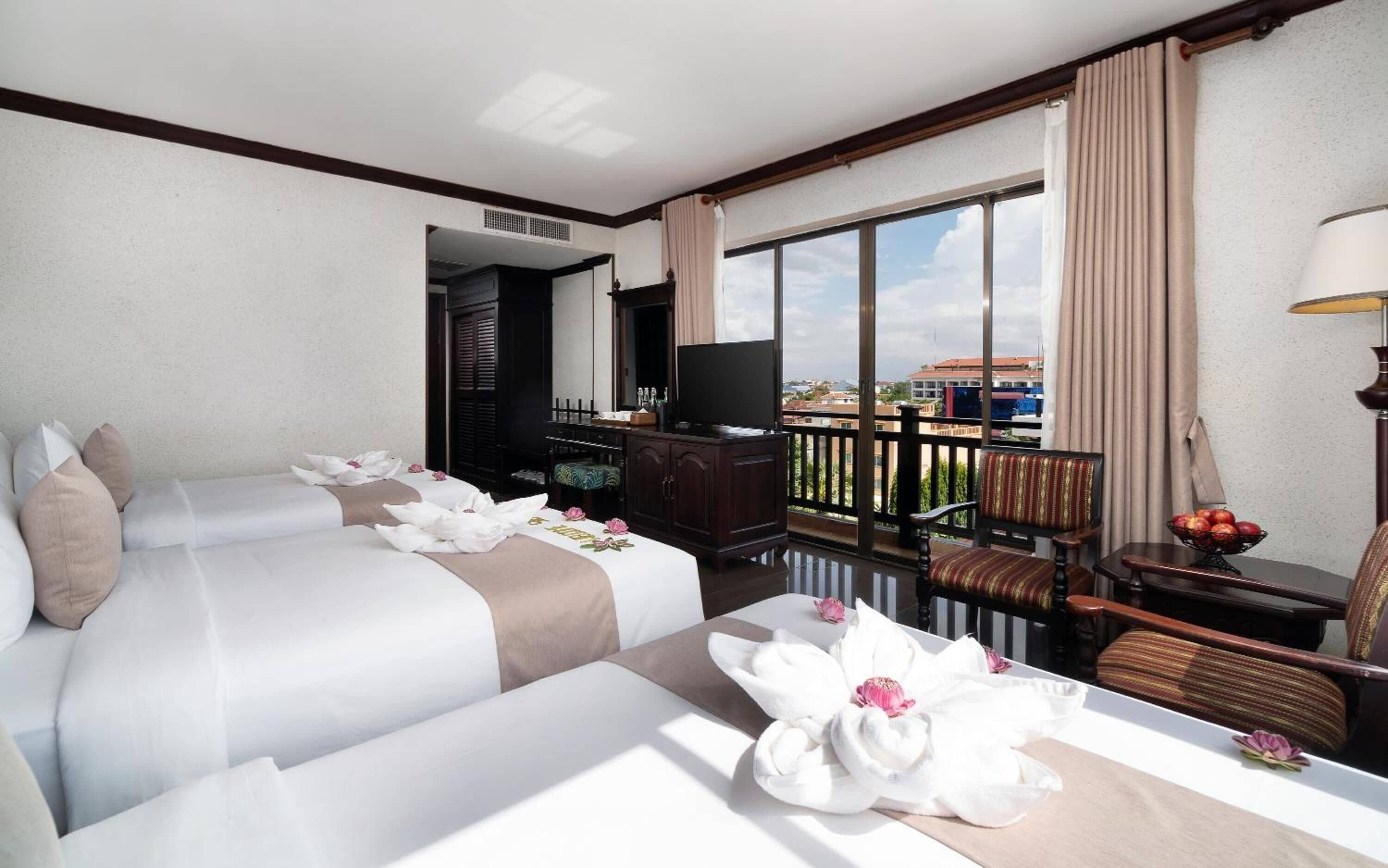 Habitación de hotel con dos camas, televisor, balcón con vistas y zona de comedor con mesa y sillas.