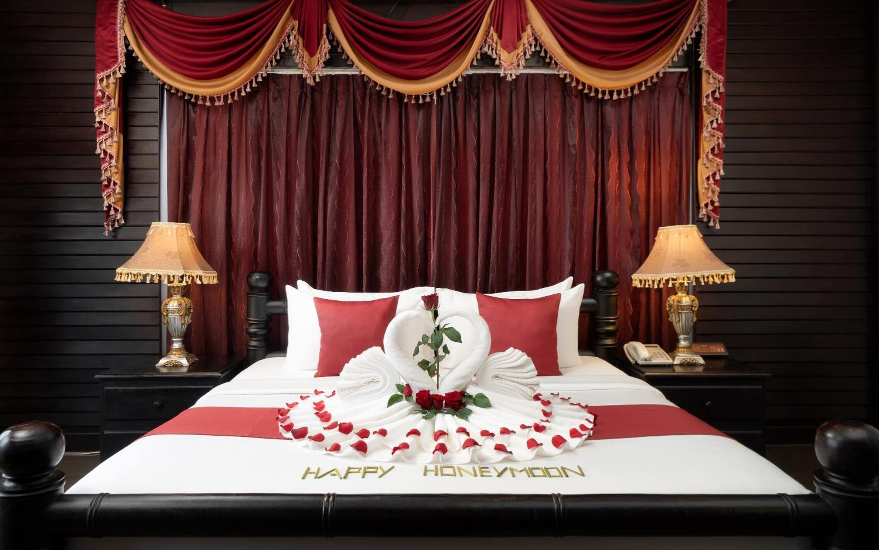 Una cama decorada con la inscripción 'Happy Honeymoon', ropa de cama roja y blanca, y un centro de mesa con una rosa.