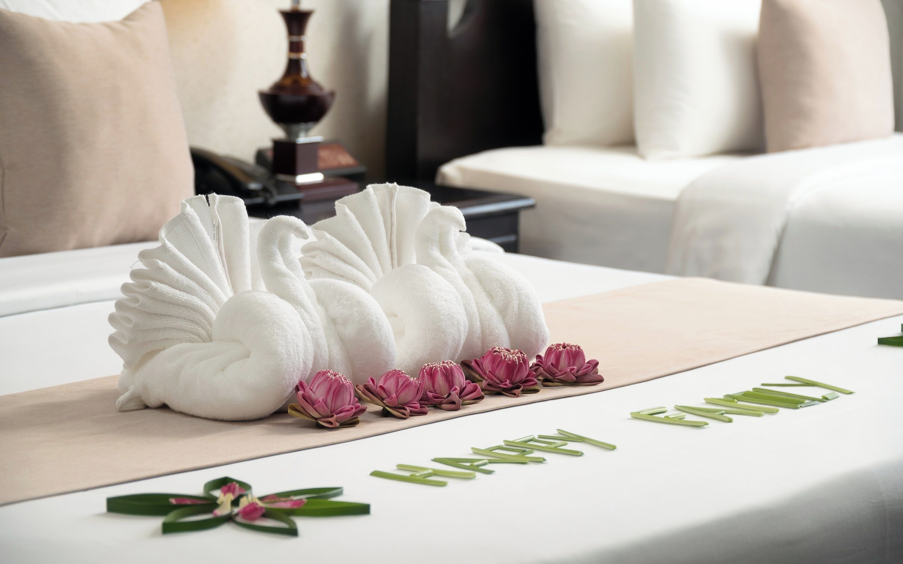 Una cama bien hecha con cubrecama blanca, toallas decorativas y flores rosadas sobre una mesa blanca.