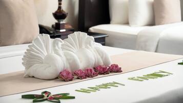 Una cama bien hecha con cubrecama blanca, toallas decorativas y flores rosadas sobre una mesa blanca.
