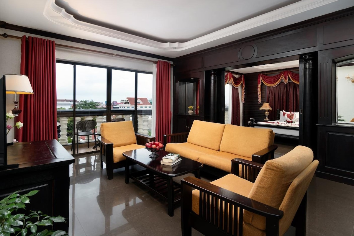 A hotel room with a sofa, armchairs, a coffee table, and a view of the city through large windows.