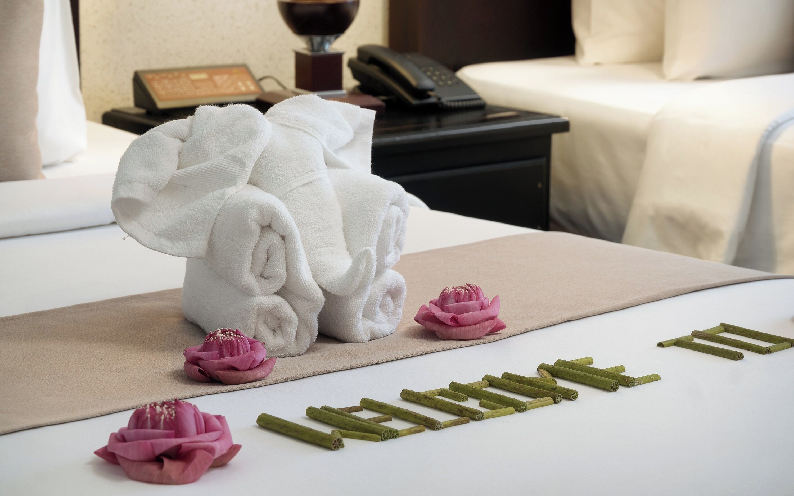 Una cama bien hecha con sábanas blancas, una mesita de noche con un teléfono y una lámpara, y una pequeña flor rosa sobre la cama.
