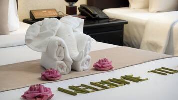 Una cama bien hecha con sábanas blancas, una mesita de noche con un teléfono y una lámpara, y una pequeña flor rosa sobre la cama.