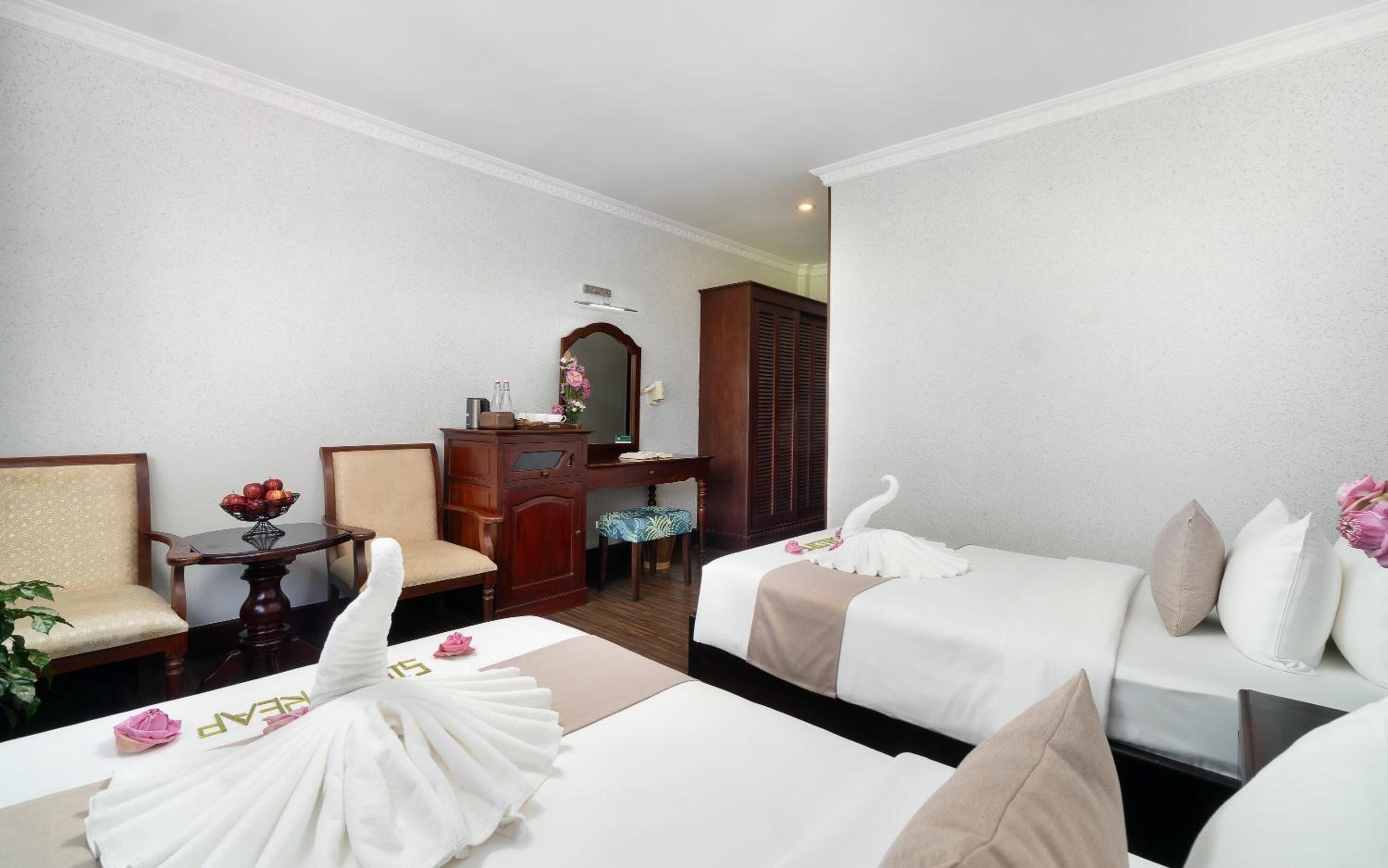 Une chambre d’hôtel avec deux lits, une commode en bois, une chaise, une petite table avec un panier de fruits et un miroir au mur.