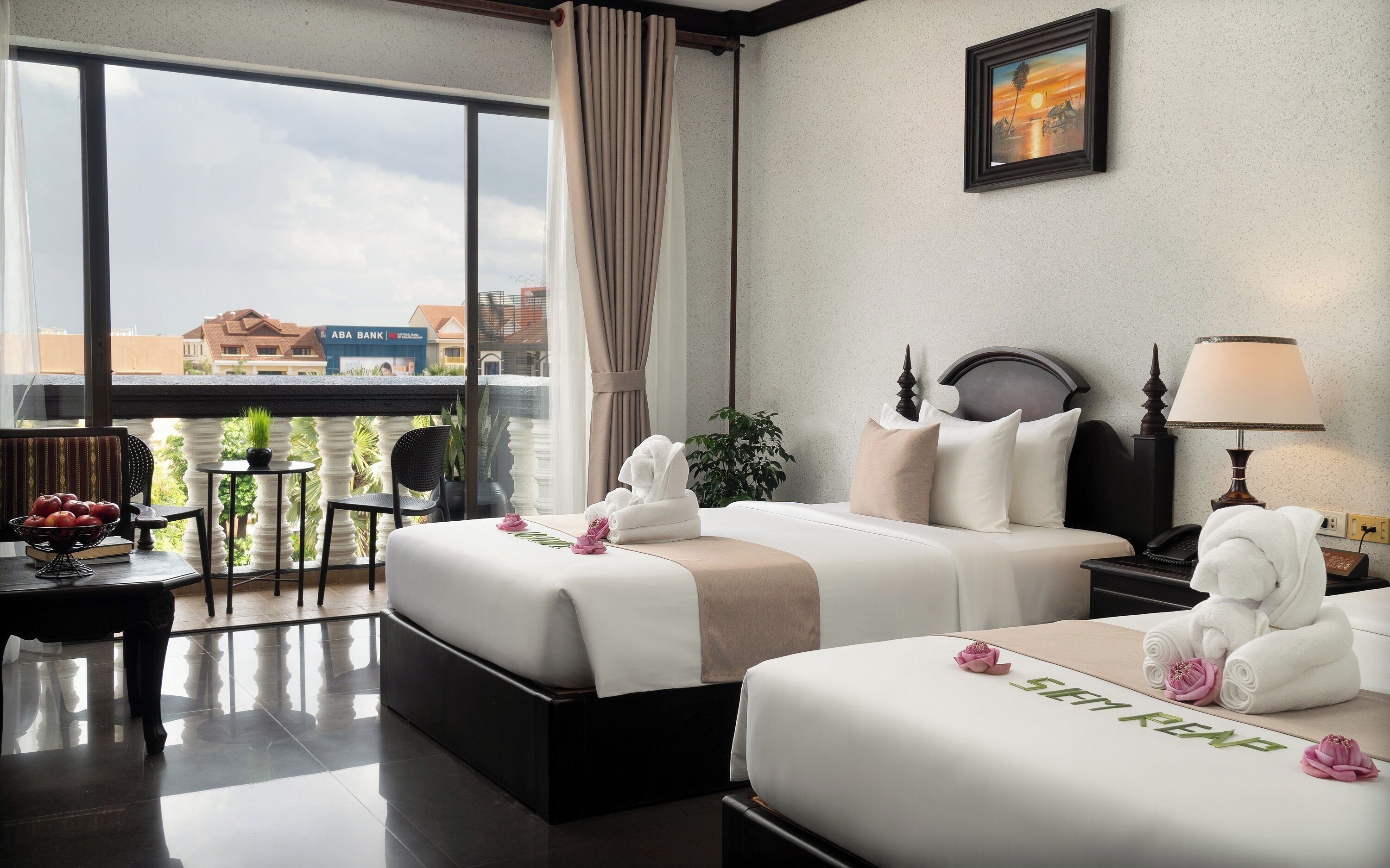 A hotel room with two beds, a view of the city, a balcony with a table and chairs, and a framed picture on the wall.