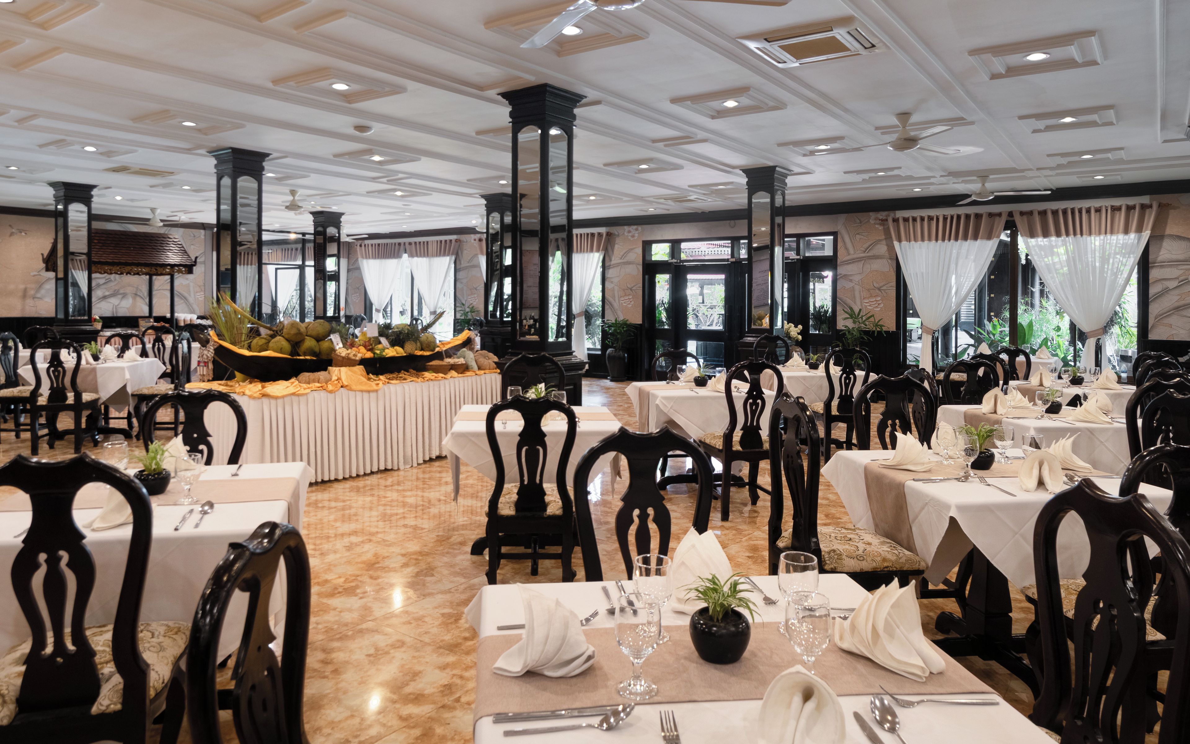 A restaurant with multiple tables, black chairs, and a buffet area with fruits and vegetables.
