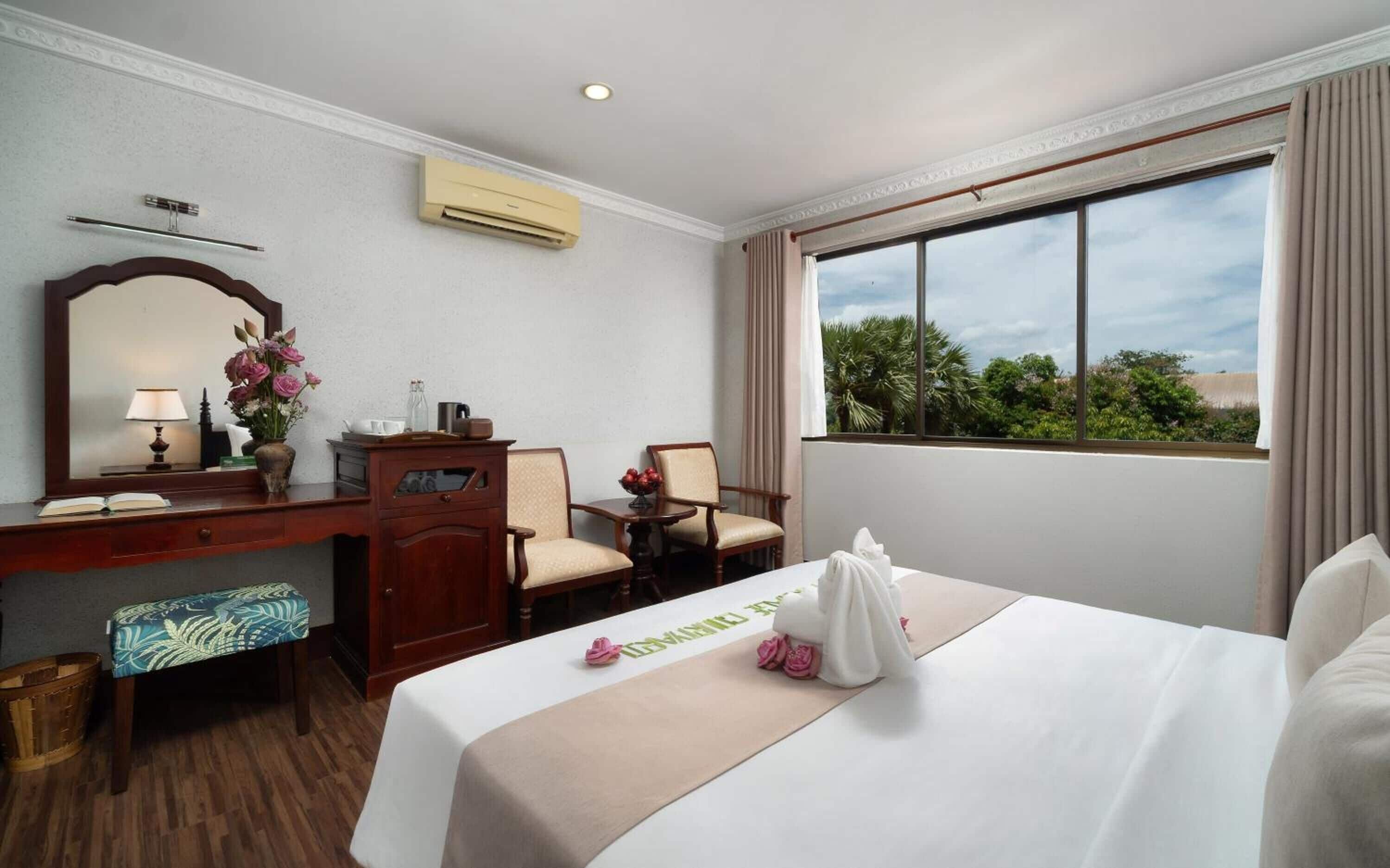 A hotel room with a bed, a desk, a chair, a mirror, and a window with a view of trees.