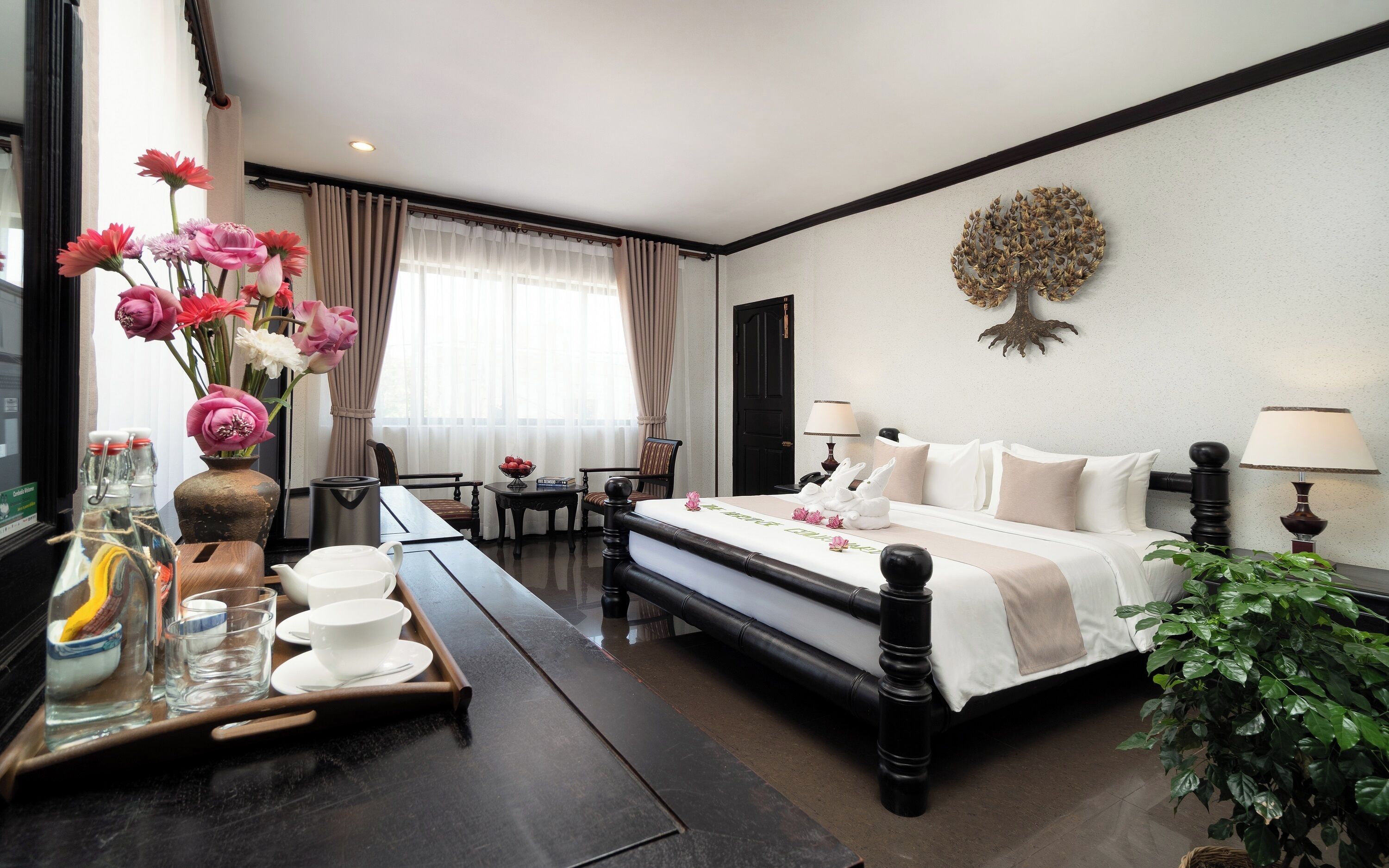 Une chambre d’hôtel avec un lit, des lampes de chevet, une table avec un plateau de verres et un vase de fleurs, une chaise, un bureau et une fenêtre avec des rideaux.
