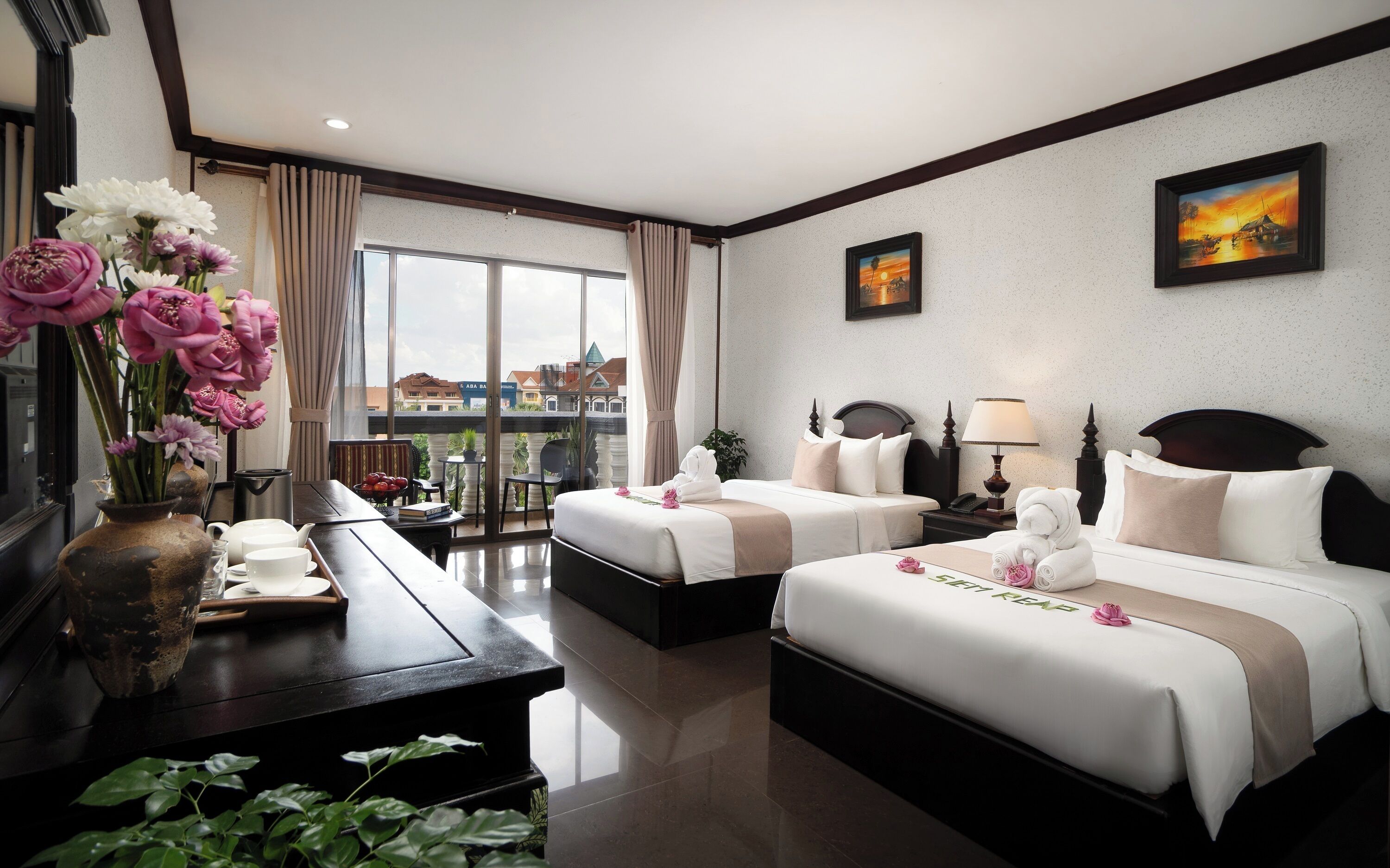 A hotel room with two beds, a desk, a vase with flowers, and a view of buildings.