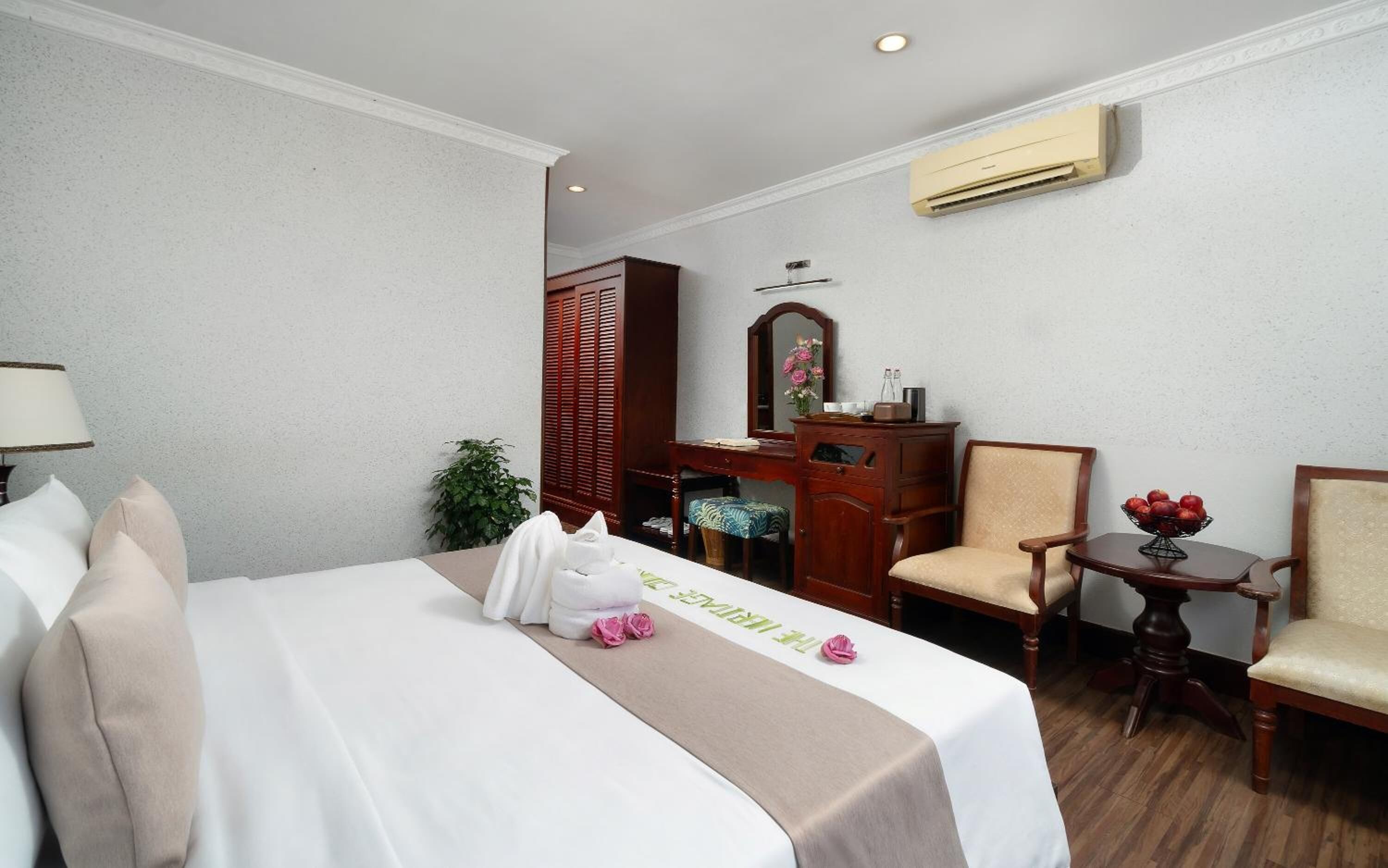 A hotel room with a bed, a desk, a chair, a small table with fruit, and an air conditioning unit.