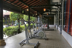 Zona de gimnasio al aire libre techada con equipo de ejercicio, piso de baldosas y vistas a la vegetación.