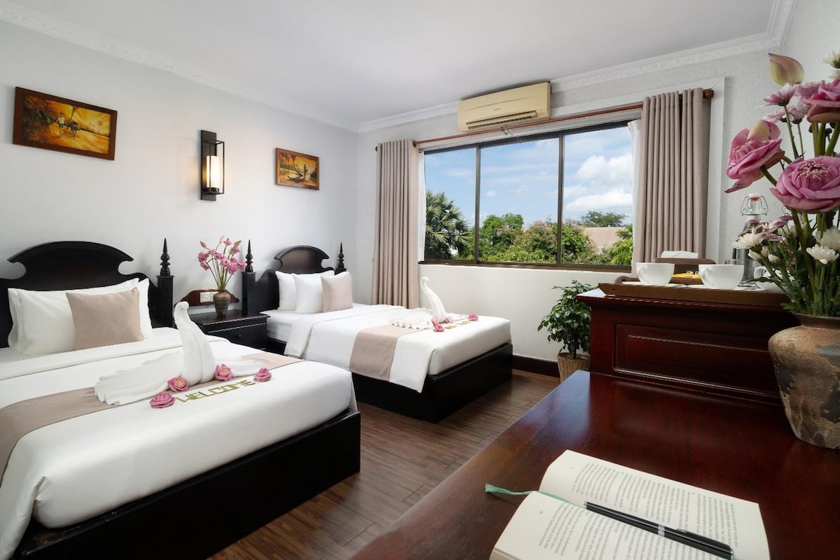A hotel room with two beds, a desk, a chair, a window with a view, and artwork on the walls.