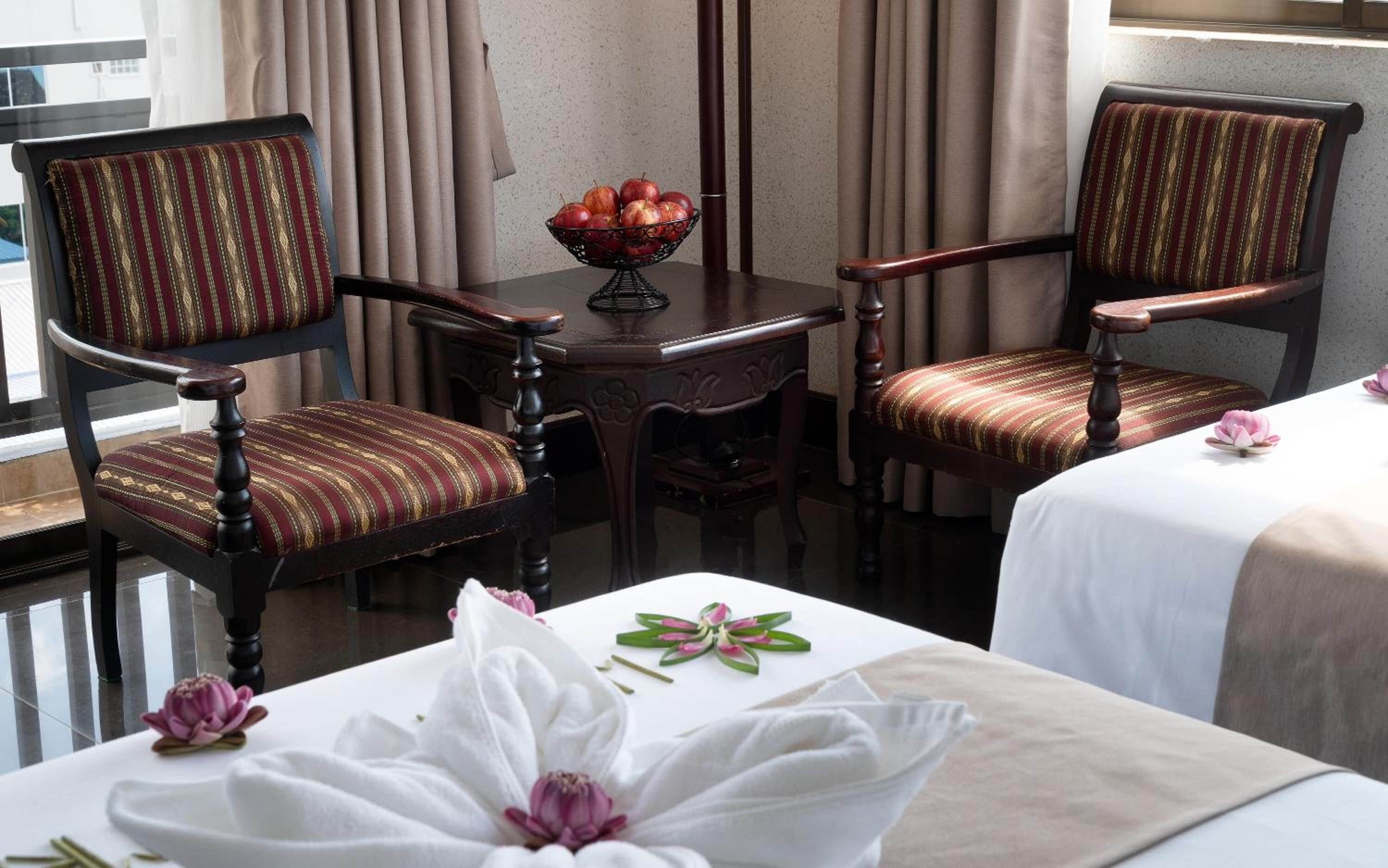 Habitación de hotel con dos sillones tapizados, una mesita con fruteros y una cama con cubrecama blanco y motivos florales.
