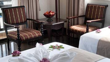 Habitación de hotel con dos sillones tapizados, una mesita con fruteros y una cama con cubrecama blanco y motivos florales.