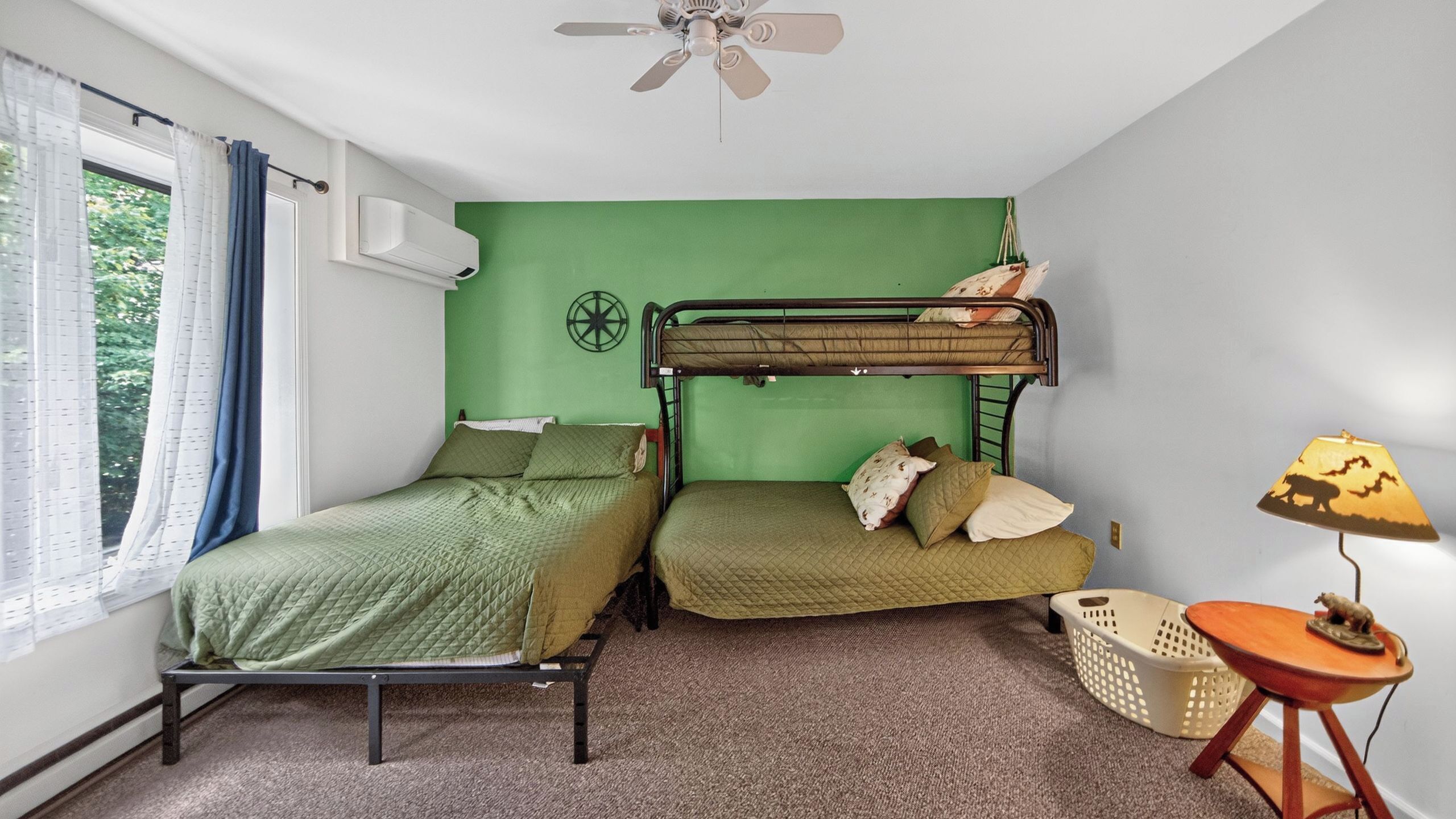 Um quarto em dormitório com beliches, um ventilador de teto, um aparelho de ar condicionado, uma janela com cortinas e uma pequena mesa com luminária.