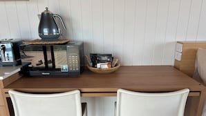 A compact kitchenette with a microwave, toaster, and coffee maker on a wooden countertop. There are two white chairs and a wooden bowl with coffee pods.