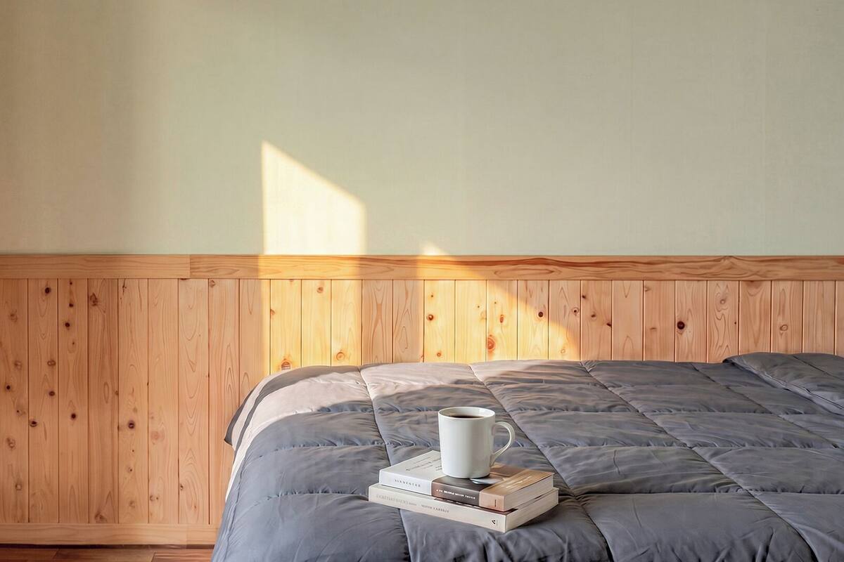 Un dormitorio con cabecera de madera, una cama con colcha gris, una taza blanca y una pila de libros.