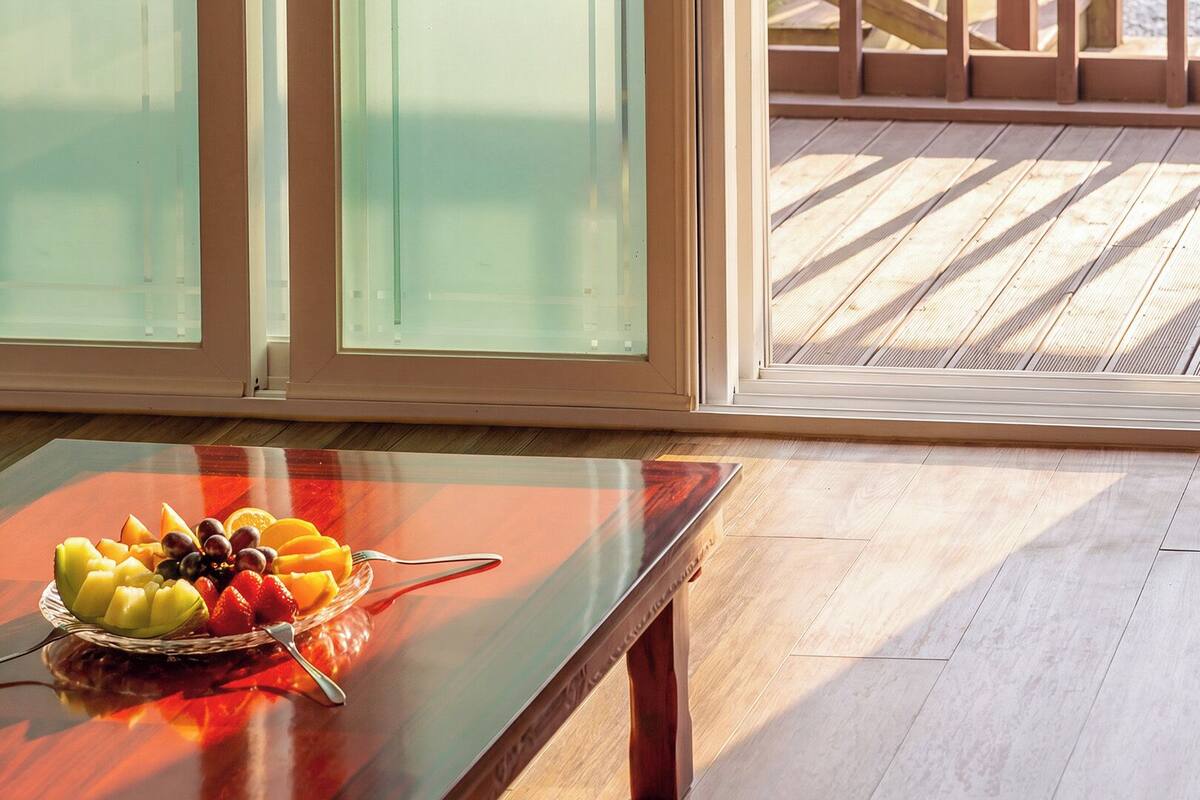 Habitación con mesa de vidrio que exhibe fruta y una cuchara, ventanas amplias y una terraza de madera afuera.