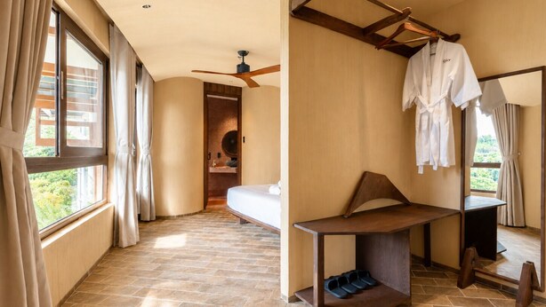 Habitación de hotel con cama, escritorio, espejo, ventilador de techo y ventana con cortinas.