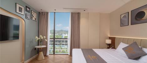 A hotel room with a bed, a TV, a window with curtains, a small table with a vase, and framed pictures on the wall.