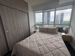 A bedroom with a bed, nightstands, a window with a view of tall buildings, and a wooden wardrobe.