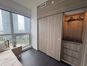 A bedroom with a large window, a bed, a nightstand, a wardrobe, and a hanging closet.