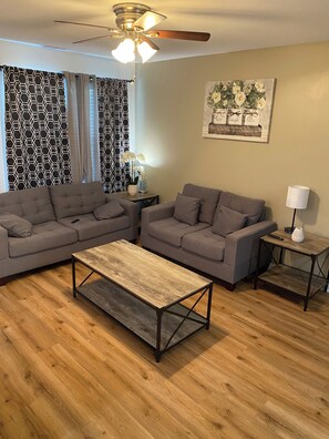 Ein Wohnzimmer mit zwei grauen Sofas, einem Holztisch, einem Deckenventilator, gemusterten Vorhängen, einem Wandbild und einer Lampe auf einem Beistelltisch.