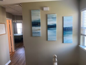 A hallway with three vertical paintings on the wall, a white sculpture, and a window with blinds.