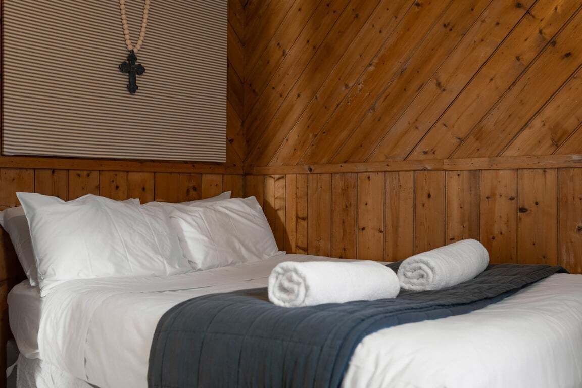 A wooden-paneled room with a bed, white bedding, and a cross pendant on the wall.