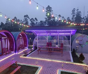 A covered outdoor seating area with tables and chairs, surrounded by tents and string lights. - Cozzy Rompok Cileunca (bandung)