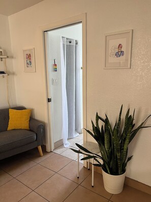 A living room with a gray sofa, a yellow pillow, a potted plant, a framed picture, and a door with a white curtain. - Las Cruces Landing Pad - Available July to November (Mesilla)