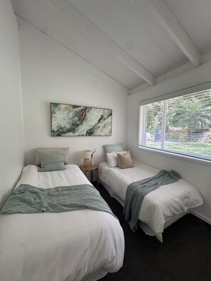 Ein Schlafzimmer mit zwei Betten, einem Fenster mit Jalousien, einem Bild an der Wand und einem kleinen Tisch mit einer Lampe.