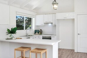 Eine moderne Küche mit weißen Schränken, einer zentralen Kochinsel mit Hockern, einem eingebauten Backofen und einem Fenster, das natürliches Licht hereinlässt.