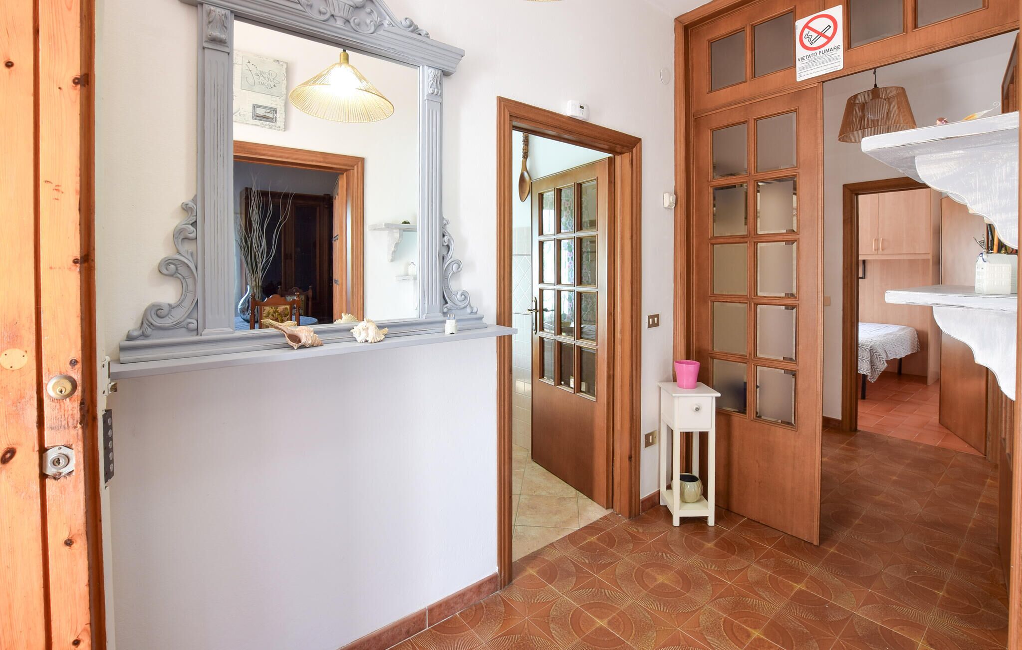 Un corridoio con una porta in legno, uno specchio, un piccolo tavolo con un contenitore rosa e un pavimento piastrellato.